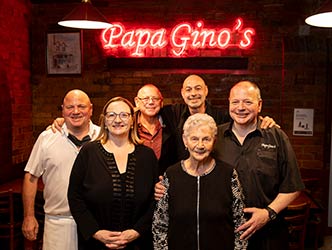 Alex Brosca and family standing in the restaurant underneath the red neon 'Papa Gino's' sign.