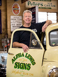 Paul Lewis standing next to a vintage vehicle with the business name 'Alpha Lewis Signs' and phone number on its door.