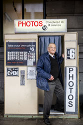 Alan Adler standing in front of a vintage photobooth with signs that reads ‘photos’ and ‘$6’ on it.