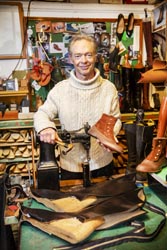 Brendan Dwyer in his studio holding a tan boot and a hammer next to a vintage sewing machine, surrounded by partly constructed pairs of shoes. 