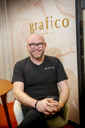 Damian Corney sitting on a chair in front of a wall with the business name ‘grafico’ on it.