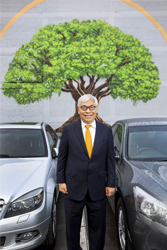 David Man Kit Yu standing between two cars parked infront of a concrete wall painted with a mural of a tree.