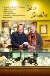 Julie and Jimmy Fong standing behind a display cabinet of jewelry, a sign on the wall reads ‘Your Jeweller’.