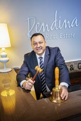 Lou Rendina standing behind a desk holding a gavel and large bell, a sign on the wall behind him reads ‘Rendina Real Estate’. 