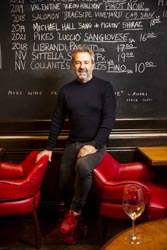 Martin Pirc leaning against a red leather chair in front of a black board with a list of wine specials written on it.