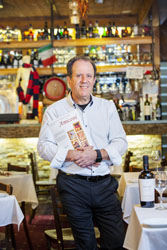 Michael Caramone standing between tables in Amiconi Restaurant holding a menu to his chest.