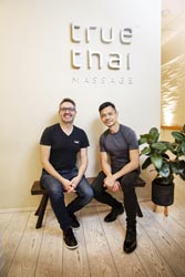Michael Wright and Tae Panuktong sitting on a bench in front of a wall with a sign that reads ‘true thai massage. 