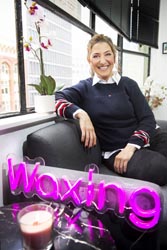 Nikki Vriends sitting on a couch in the Zip Zip Wax reception area near a scented candle and a hot pink neon sign that reads ‘waxing’.