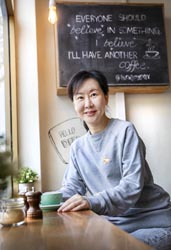 Rongrong Wang sitting at a table with a cup of coffee in front of a sign that reads ‘everyone should believe in something and I believe I’ll have another coffee’.