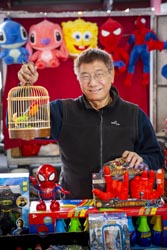 Yik Muoa Hong standing behind the counter of Motion City Development, holding onto a toy birdcage, surrounded by other toys.