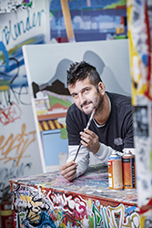 Man with paint bushes and spray cans with art in background