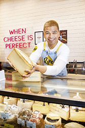 Smiling man behind counter holding large wheel of cheese