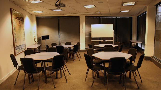 Room with four hexagonal tables, each with six chairs; lectern and whiteboard 