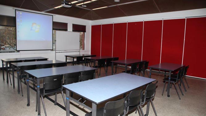 Room with red wall and eight tables each with three chairs facing a projector screen and whiteboard