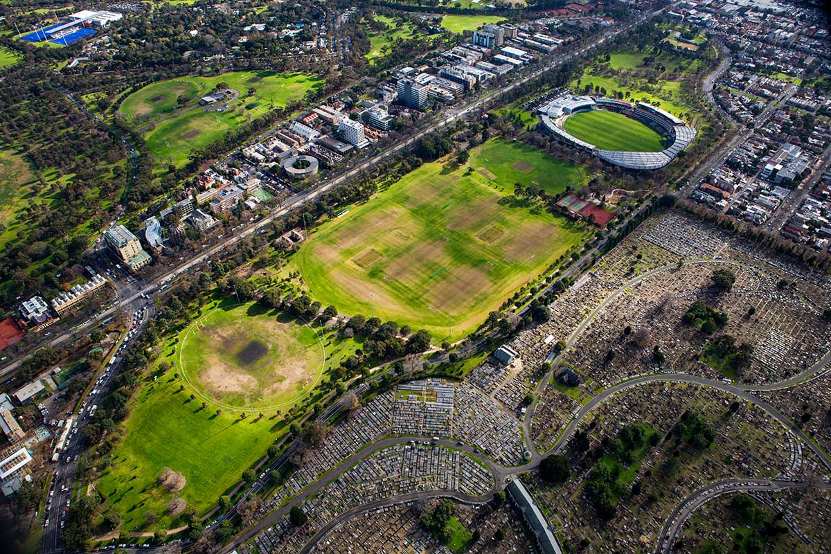 An aerial view of Princes Park featuring large sports fields, separated by avenues of large trees. Royal Parade and the cemetery can be seen outside the park's boundary.