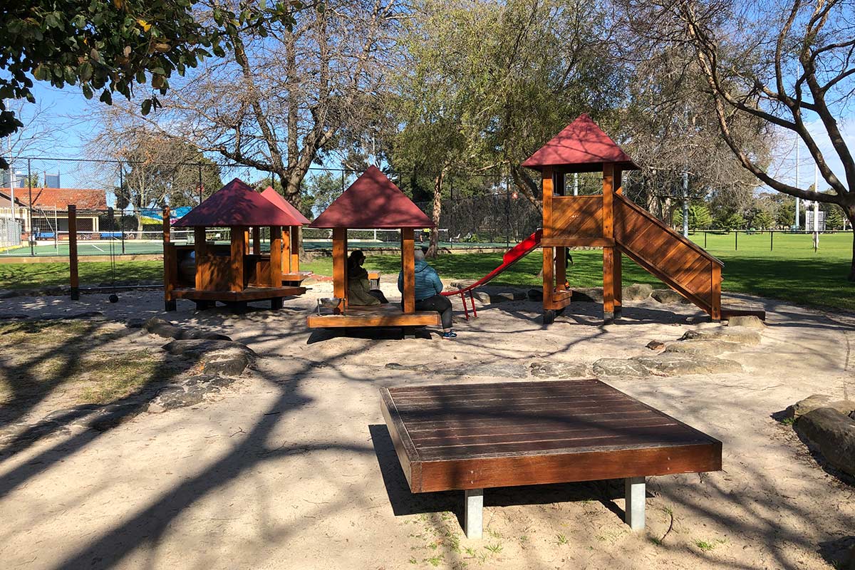 Playground for infants with small wooden structures and a slide, each with red triangular rooftops.