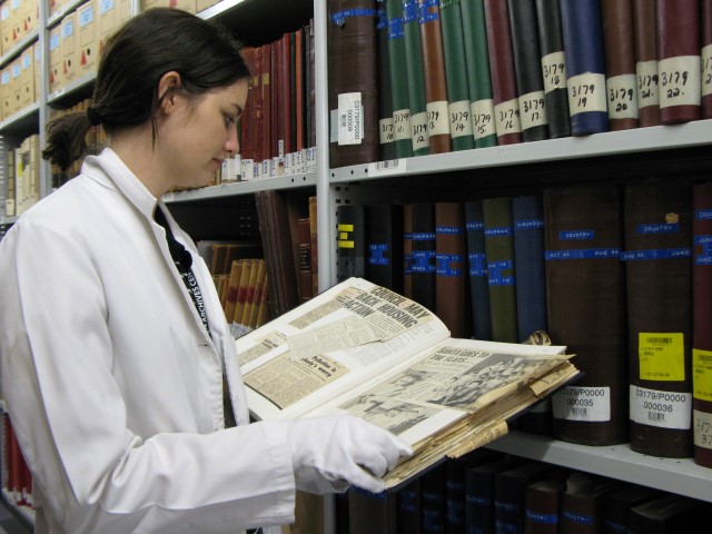Women in white coat and glovesholding book containing newspaper clippings 