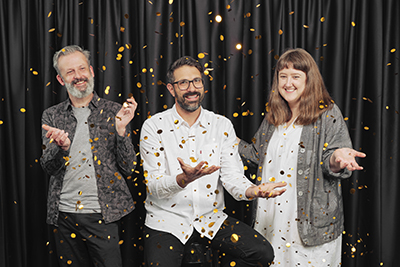 Three people standing in front of a velvet curtain throwing confetti in the air