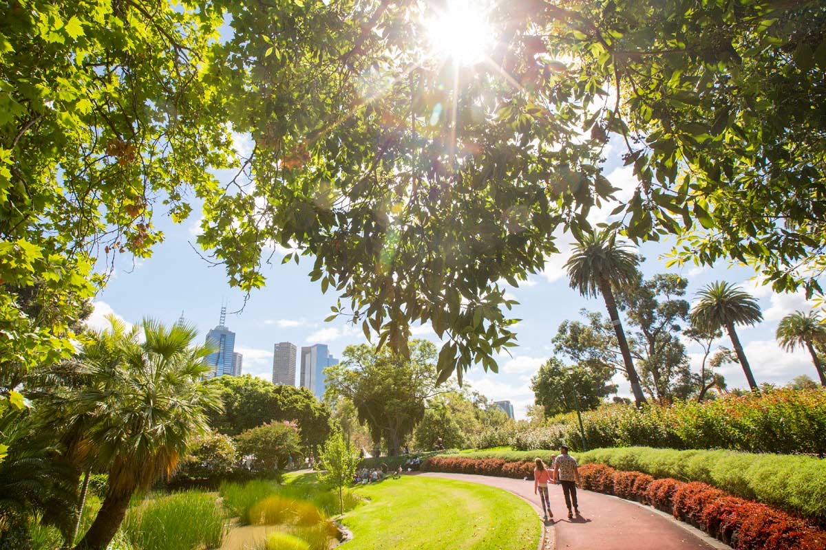 People walking on a curved path in the gardens heading towards the city. The view is framed by overhanging foliage with the sun shining through.