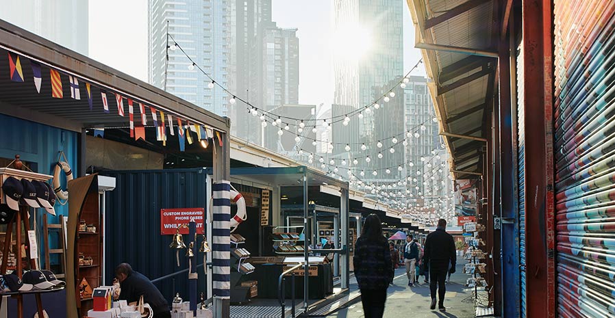 People walk alongside market stalls. City high-rises can be seen nearby.