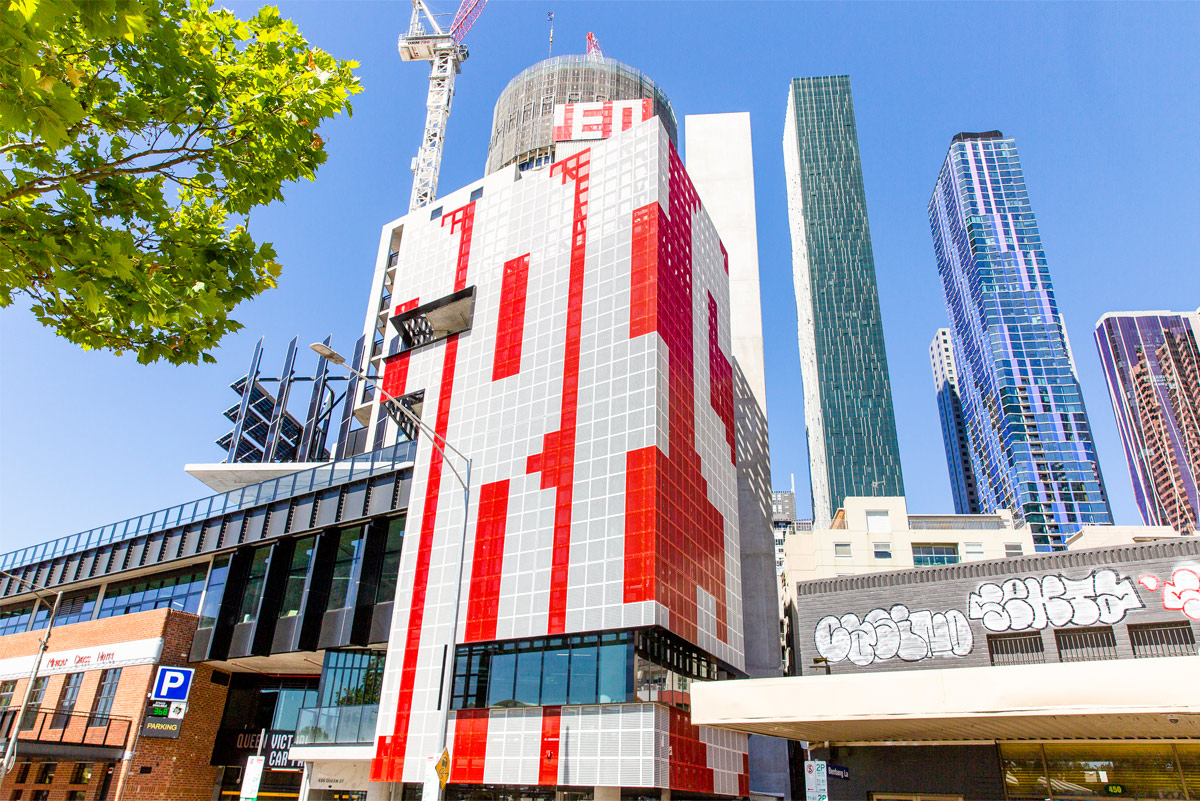 View of the red and white perforated metal screens on two sides of a building, and cladding the top of a taller building in the background.