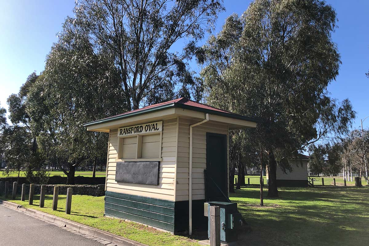 The scorebox, on the edge of the oval and next to the road, is a tiny wooden building with dark green and cream weatherboards, red roof, shuttered windows and steps leading to the door.
