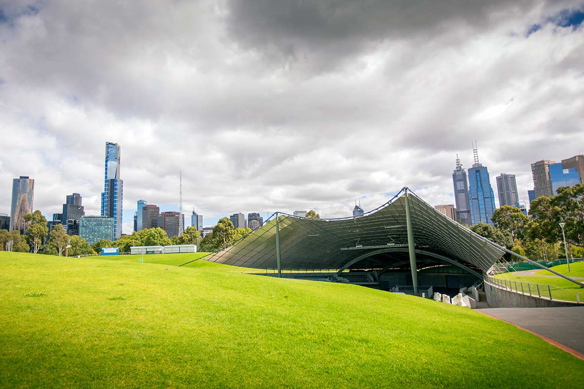 The Sidney Myer Music Bowl outdoor concert venue under a canopy. There is a large grassy hill in the foreground and city buildings in the background.