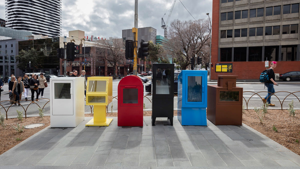Large public artwork, consisting of a row of brightly coloured newspaper stands, next to a major road.