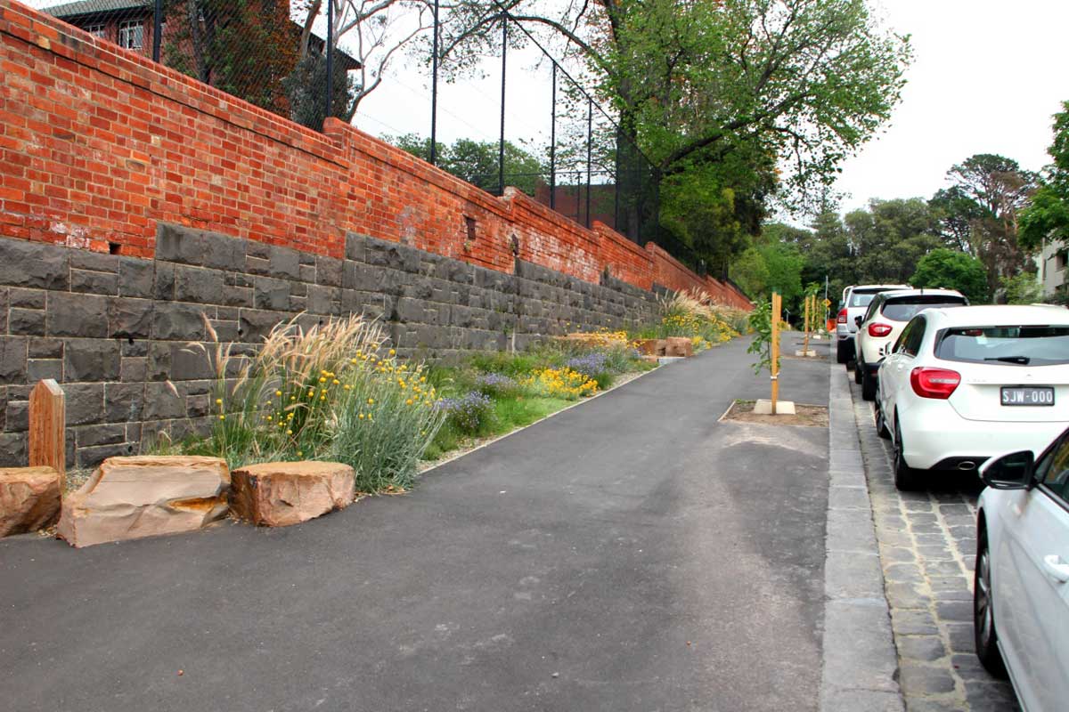 Streetscape planting of various flowering plants, groundcovers and grasses. The plantings runs along a brick wall on a slight uphill slope, and also alongside a wide footpath.