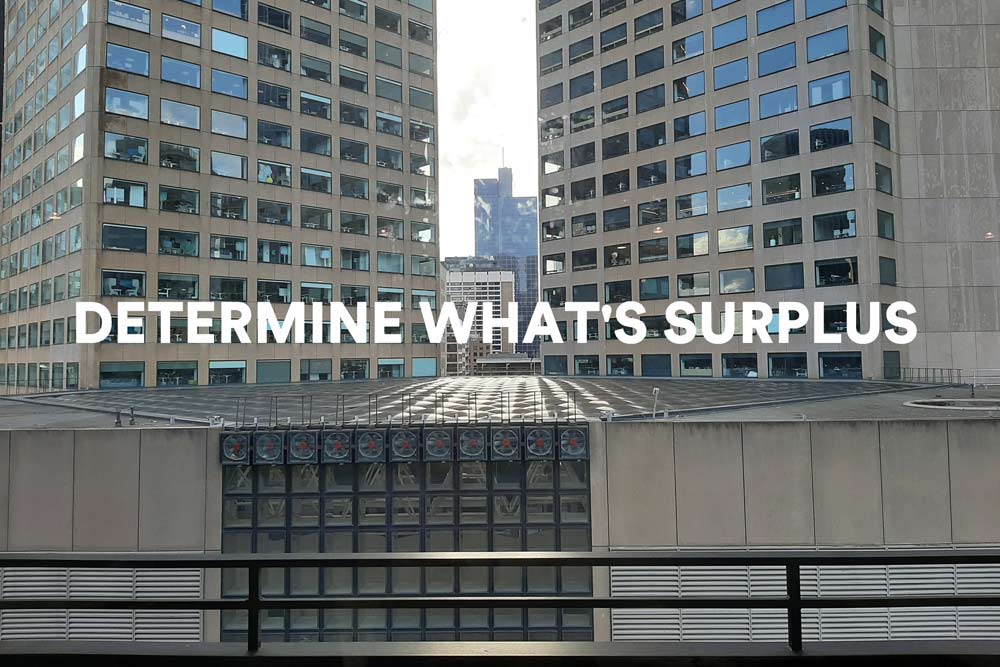 The words 'DETERMINE WHAT'S SURPLUS'  in large white text, against a background showing closely cropped view of large concrete office buildings.