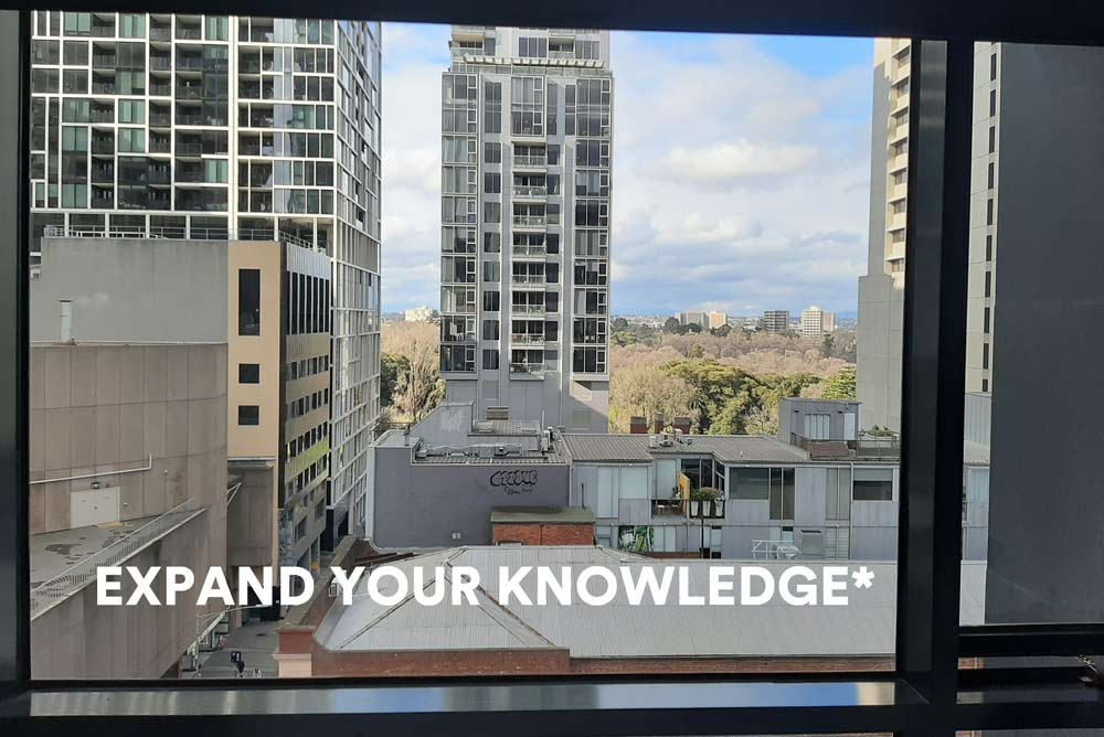 The words 'EXPAND YOUR KNOWLEDGE*'  in large white text, against a background view through a window showing high-rise buildings and rooftops and sky, buildings and trees in the distance.