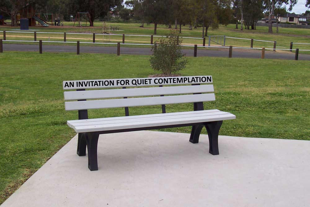 A white park bench with the words 'AN INVITATION FOR QUIET CONTMEPLATION' shown across one of the planks that forms the backrest. The bench is on a plain section of pavement and surrounded by green parkland.
