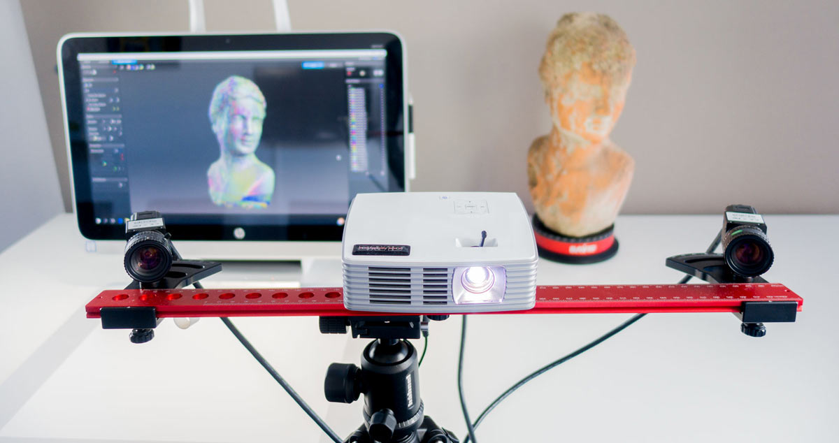 A projector in front of a plaster bust on a desk, with an image of the same bust shown on an adjacent computer screen