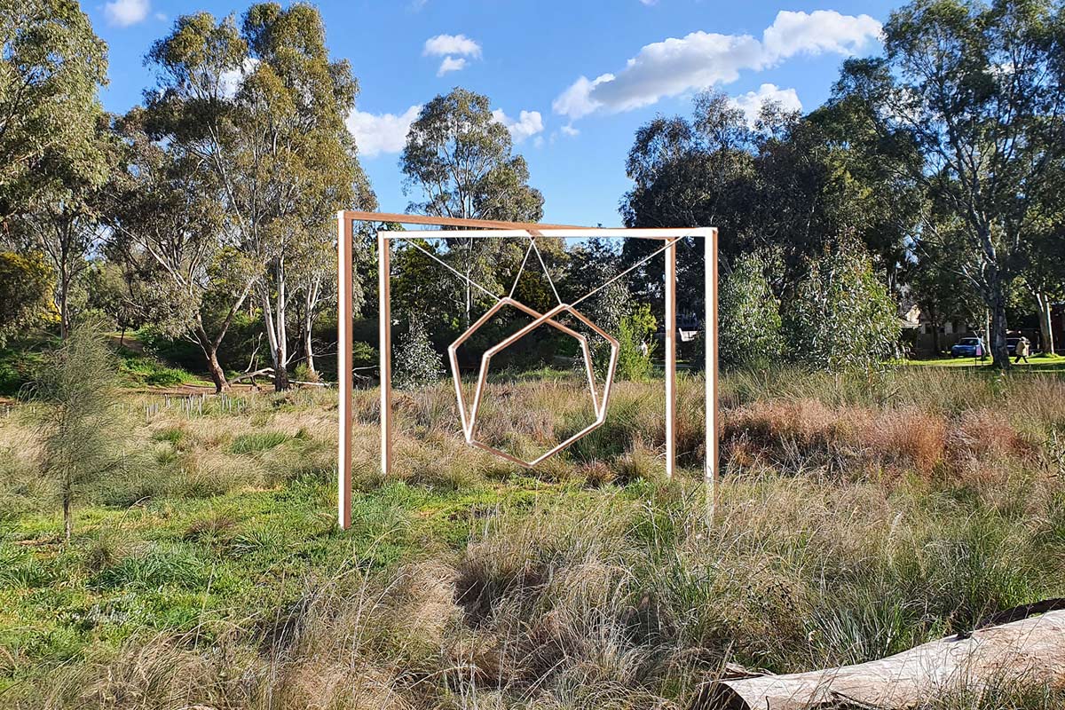 The sculpture of hexagonal frames, suspended from a cross frame structure, shown in a park among long grass, logs and gum trees.