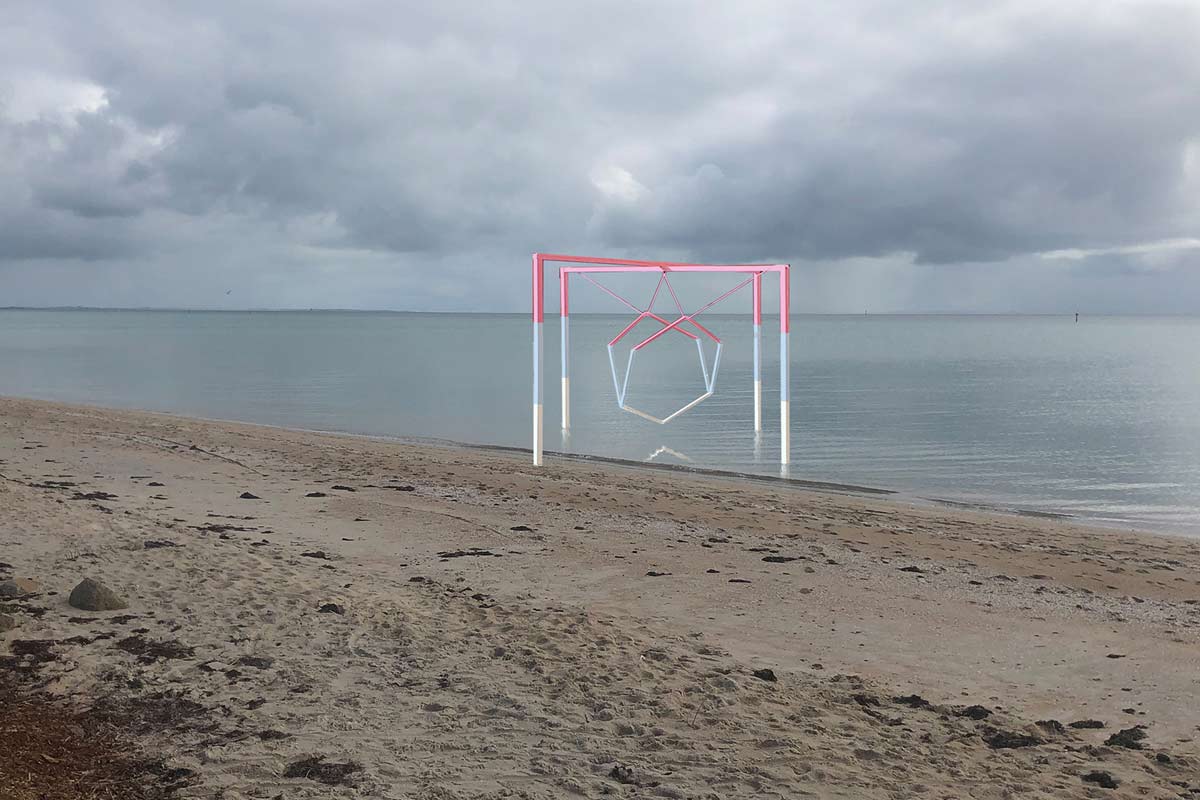 The sculpture of hexagonal frames, suspended from a cross frame structure. The bottom section of the structure/hexagons is white, the middle is pale blue and the upper section is pink. It sits on a beach, straddling the shoreline, under a cloudy sky.