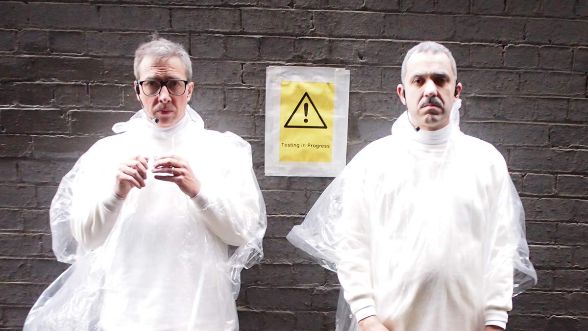 The artists wearing white clothes, plastic ponchos and microphone headsets, standing in front of a brick wall. On the wall is a sign that reads '! Testing in Progress'