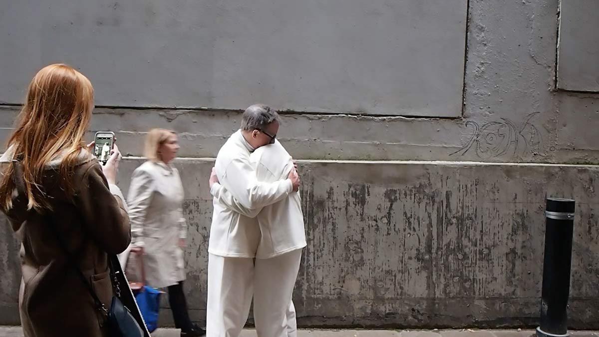 The artists hugging on another on a city street. A woman is walking past and another is taking a photo of them.