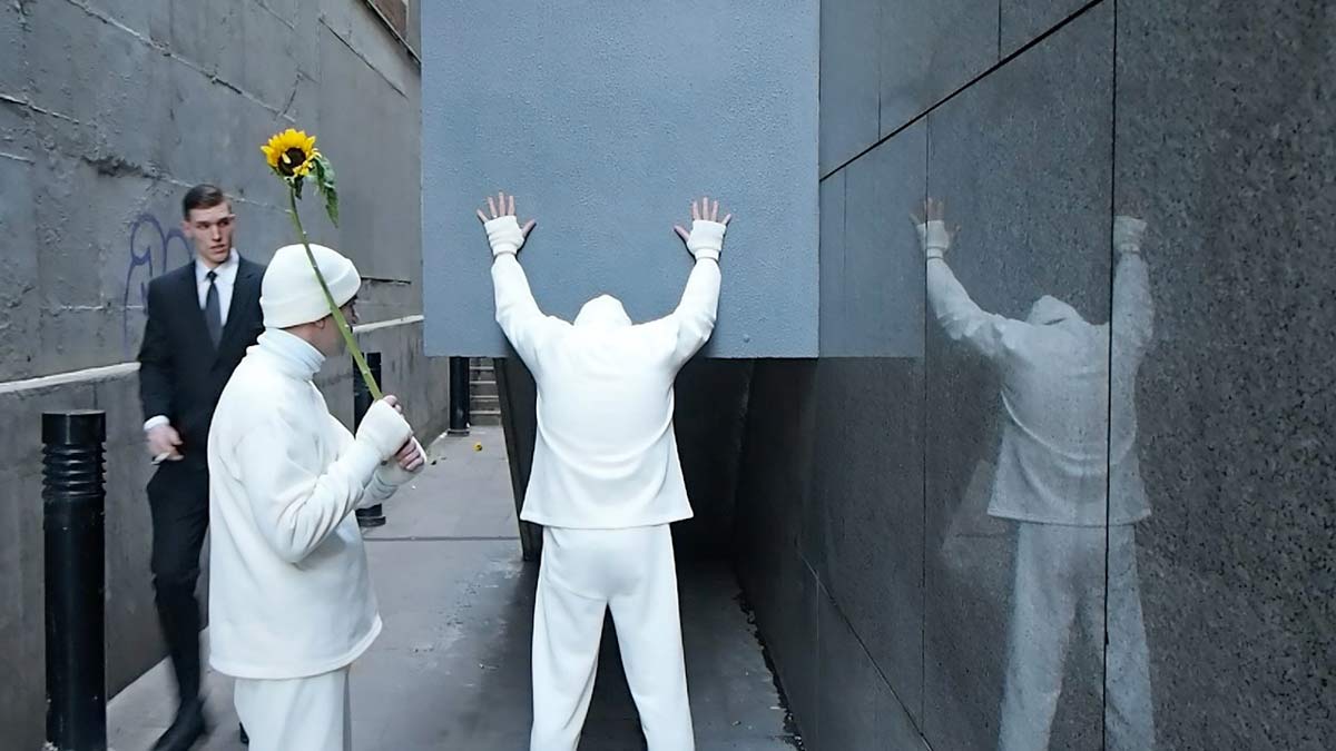 One artist stands facing a wall with his arms up, as though about to get hit; the other artist stands behind him holding a sunflower like a cricket bat. A passer-by glances at them