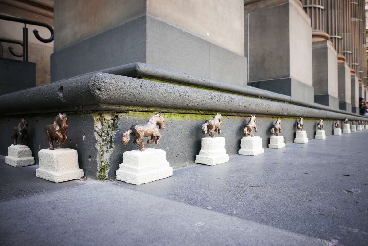 Small sculptures of bronze-coloured horses mid-trot on white plinths. More than a dozen of the sculptures are arranged facing forwards and evenly spaced around the edge of a building and around a corner