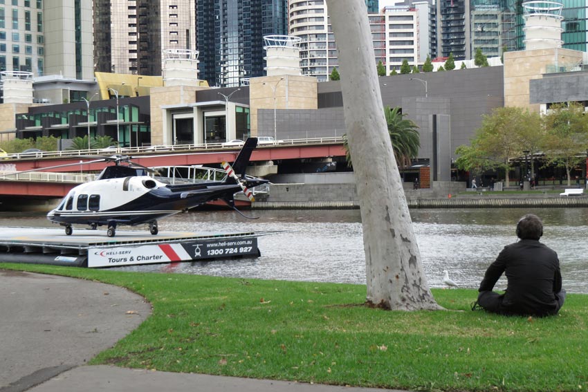 The artist, wearing headphones and sitting on the grass in Batman Park, next to the river and helicopter on a helipad