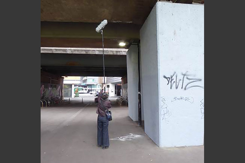 The artist standing underneath a concrete overpass, holding up recording equipment attached to a long pole