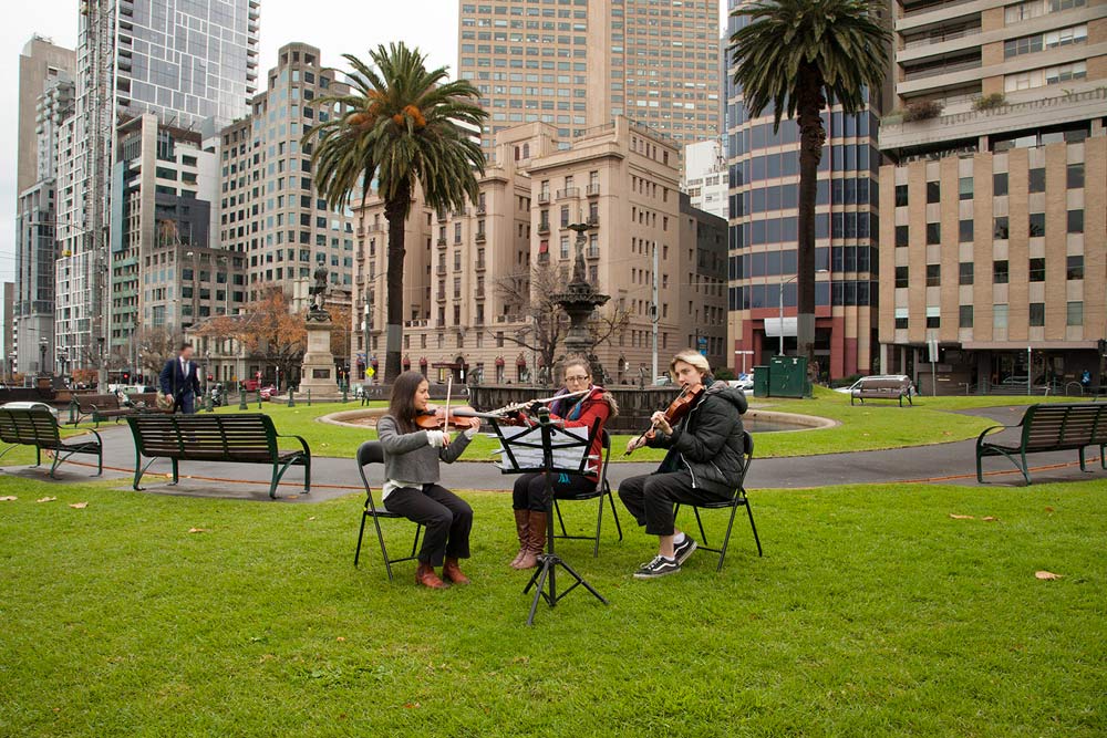 Two violinists and flautist seated in behind a music stand in a public green space, playing their instruments