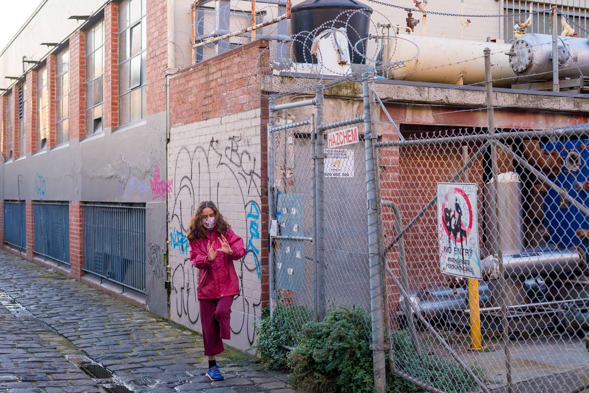 A person dressed in pink and wearing a face mask, posed standing on one leg and with their hands outstretched. They are in a cobbled laneway, next to a graffiti covered wall and wire fencing with Hazchem / No entry signs