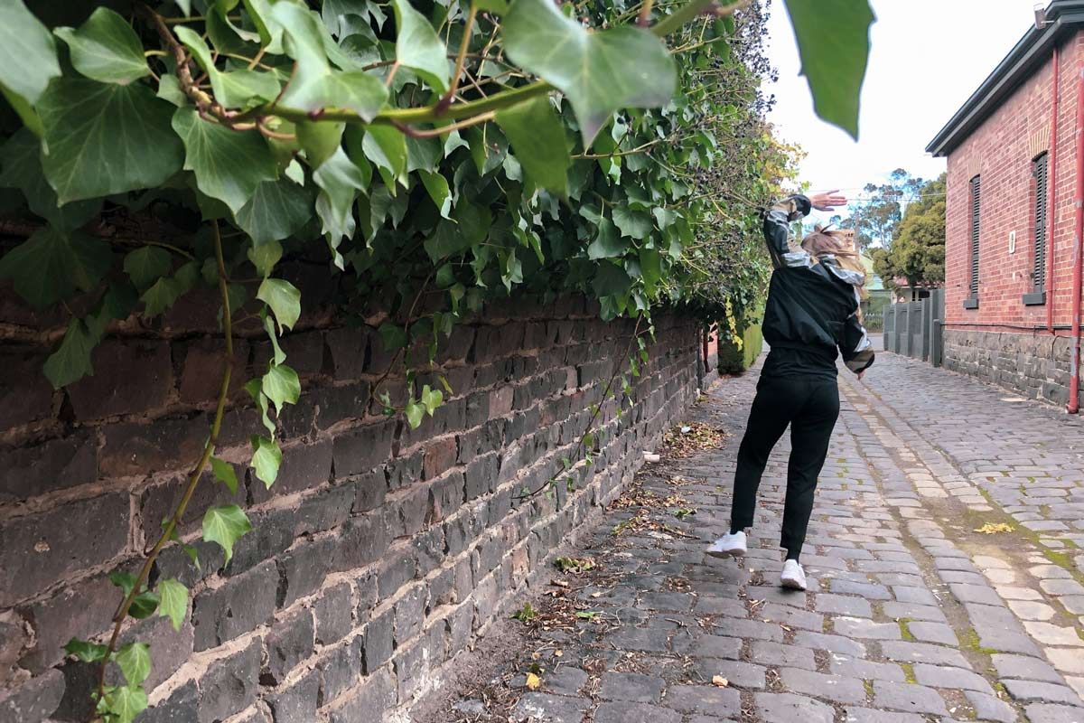Back view of a person dressed in black, mid-stride and with one arm held above their head. They are in a narrow cobblestone laneway, next to a wall half-covered by ivy.
