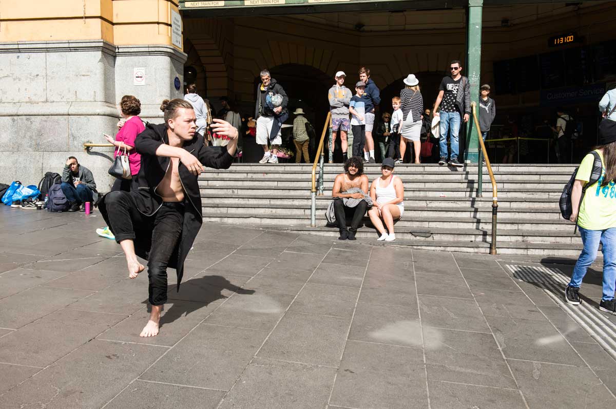 A barefoot man wearing black trousers and an opn black coatm performing dance moves in front of Flinders street station. He is being watched by the artist and other bystanders