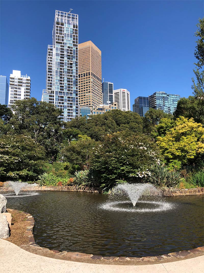 Ornamental lake in Treasury Gardens with fountains, surrounded by trees and shrubs, with CBD buildings nearby in the background.