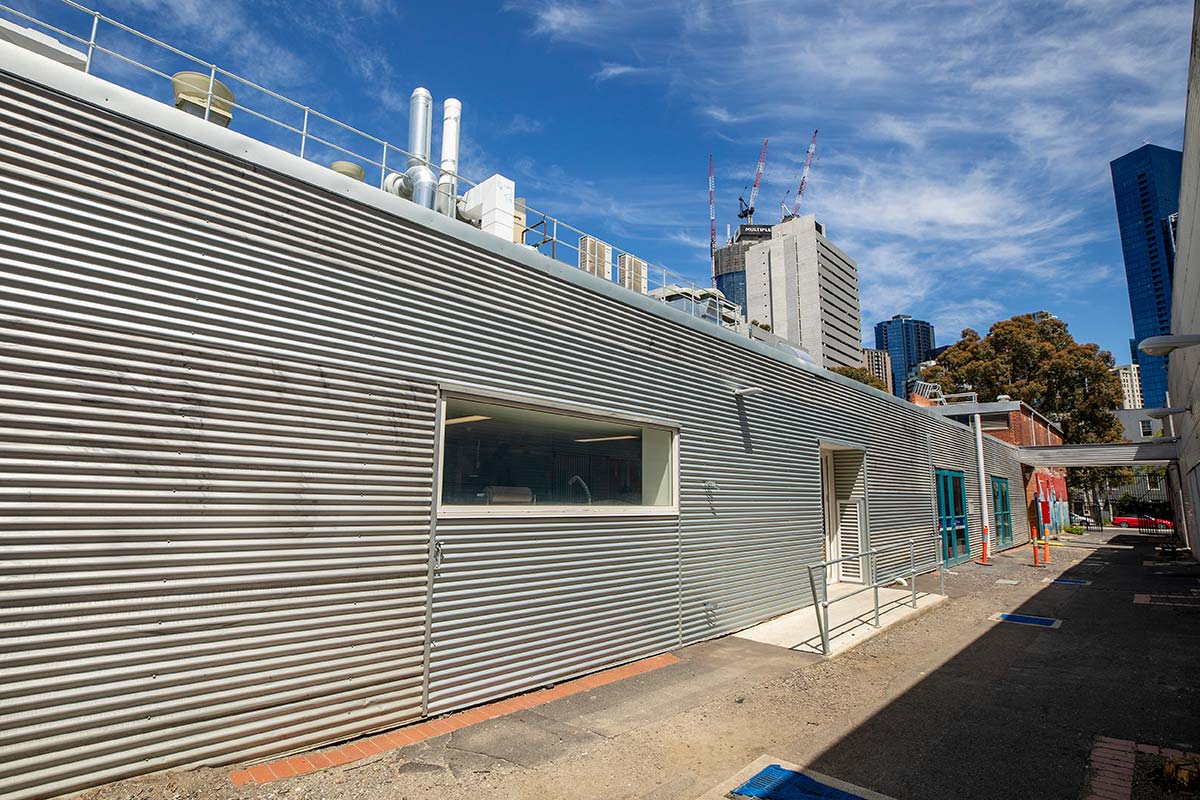 The laneway before greening works. The one-storey buildings adjacent to the path are clad in silver corrugated metal. The path's surface is part gravel and has some cracked concrete paving, with no greenery and partly shaded by the building on one side.