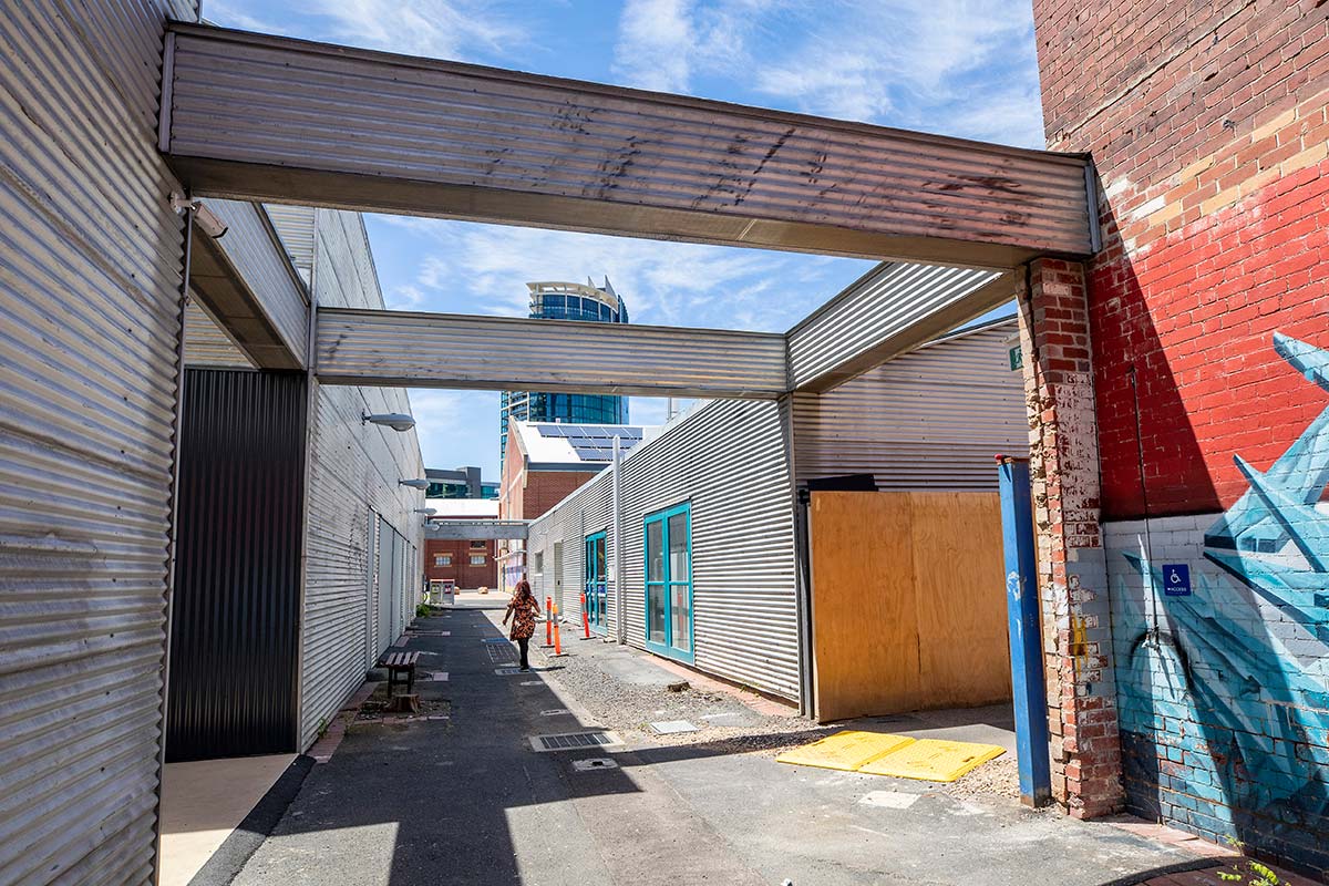 A section of the laneway with several metal gantries spanning the width of the lane. A section of brick wall is painted with a mural and the path is uneven with a mix of paved areas, bitumen and gravel.