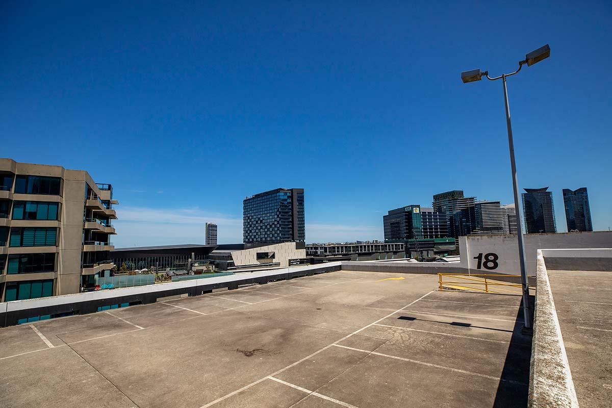 Empty rooftop paved with concrete and with parking spaces marked in white. Tall city buildings can be seen nearby.