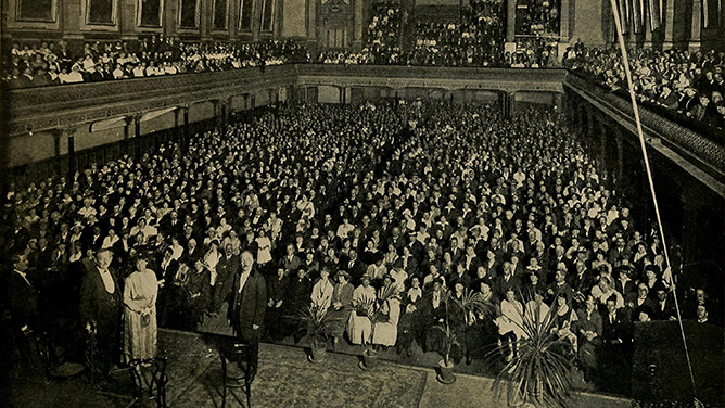 A black and white photograph of a very crowded hall with people standing on a stage